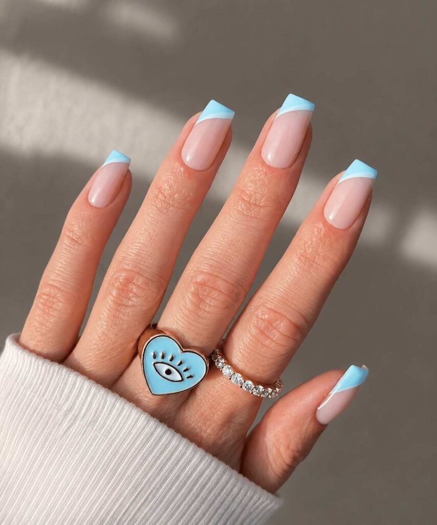 French nail manicure includes a clean pale nude pink gel base applied across the nail bed to enhance natural finger appearance, the soft sky blue or baby blue shade add a refreshing colorful pop, and the asymmetric diagonal slant stripes of painted sky-blue to one side of tips.