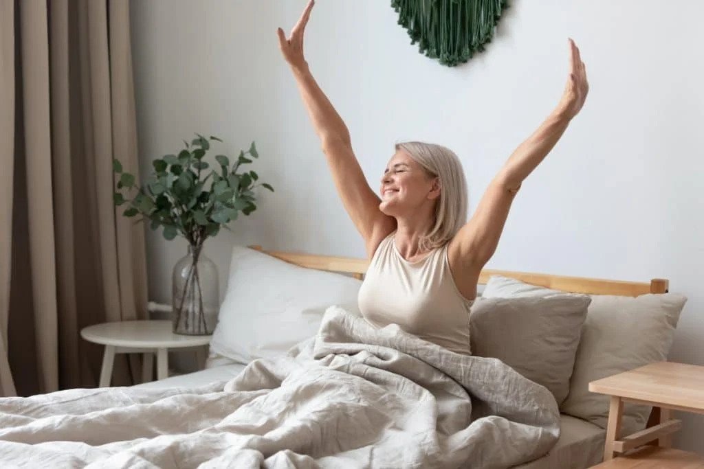 How Can You Integrate Mindfulness for Anxiety into Your Daily Morning Routine? A cheerful, mature woman is pictured waking up refreshed and stretching her arms in bed, enjoying a cozy, neutral-toned bedroom setting.