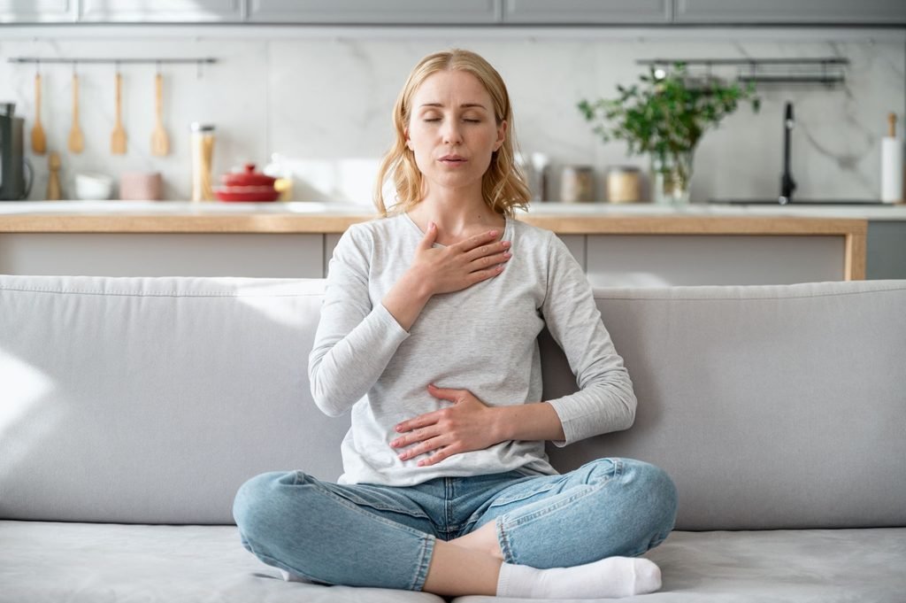 Minute 2: Slowly Breathe With Intention --A woman is sitting on a sofa with their eyes closed, placing one hand on their chest and the other on their abdomen while practicing a deep breathing or relaxation exercise