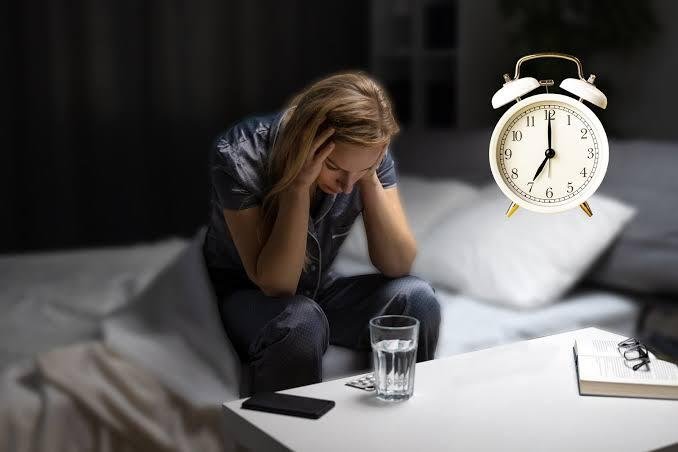 The 5 Minute Morning Routine for Anxious People - A woman sitting on the edge of her bed in a dark room, holding her head in her hands with an anxious expression while a large white alarm clock floats nearby showing 7:00
